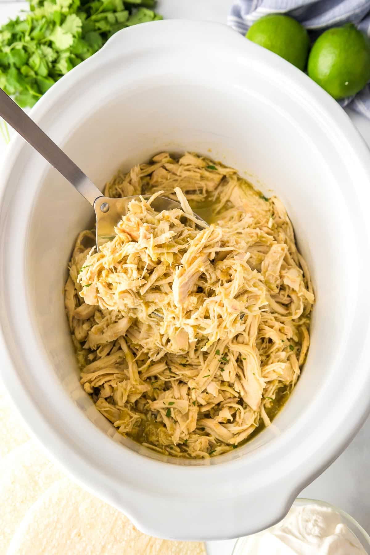 Shredded chicken in a slow cooker with a serving spoon, surrounded by fresh cilantro and limes.