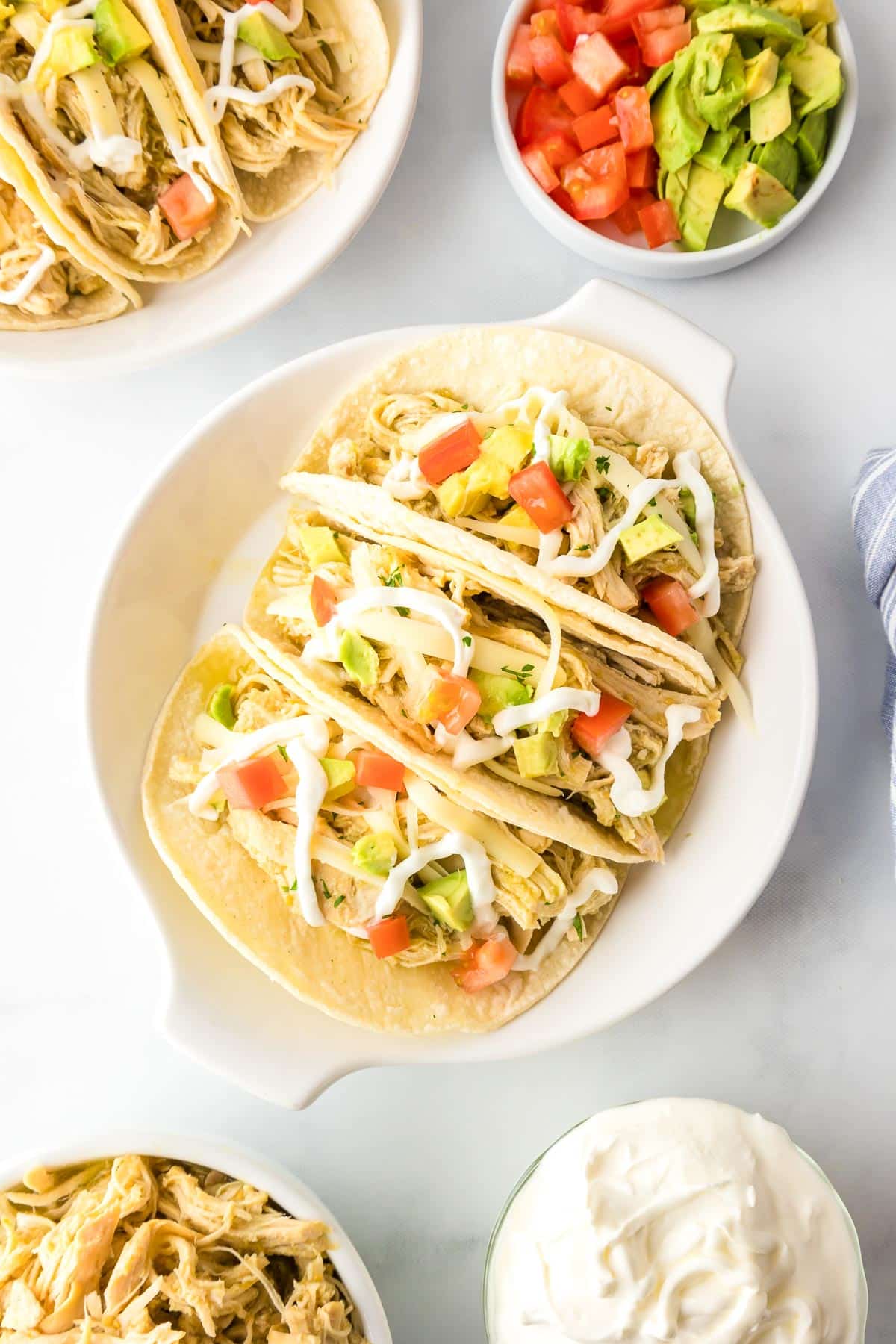 Tacos filled with shredded salsa verde chicken, diced tomatoes, diced avocados, and drizzled sour cream on a plate, surrounded by bowls of ingredients on a counter.