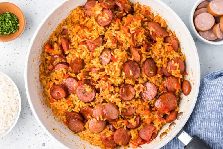 A skillet filled with cooked rice, sliced cooked smoked sausage, and red bell peppers, with a bowl of white rice and a bowl of sliced sausage nearby.