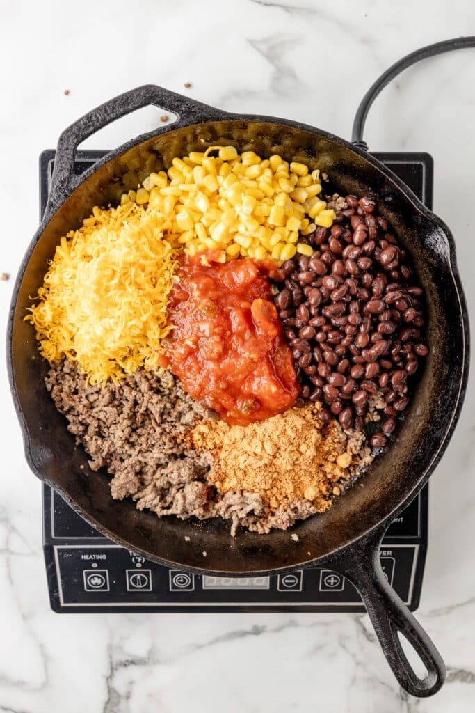 A cast iron skillet on an electric burner full of cooked ground beef, shredded cheese, corn, salsa, black beans, and taco seasoning before mixing..