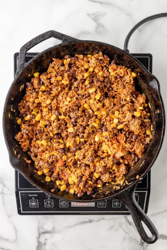 A cast iron skillet on a stovetop, filled with the base ground meat, black beans, corn, and diced tomatoes.