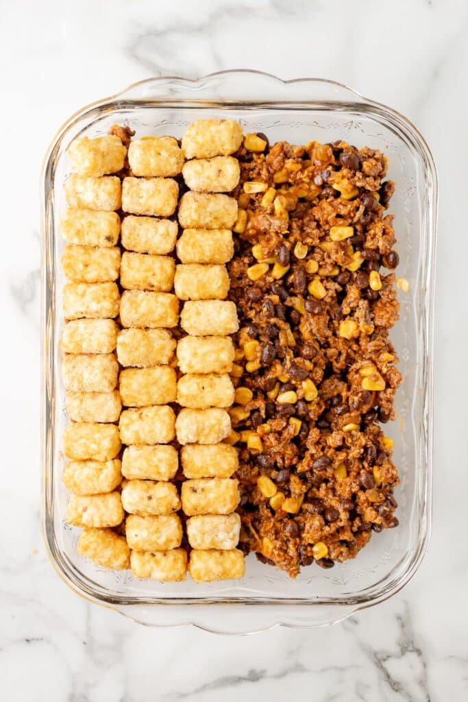 A glass baking dish full of taco casserole filling filled halfway with three lines of tatertots.