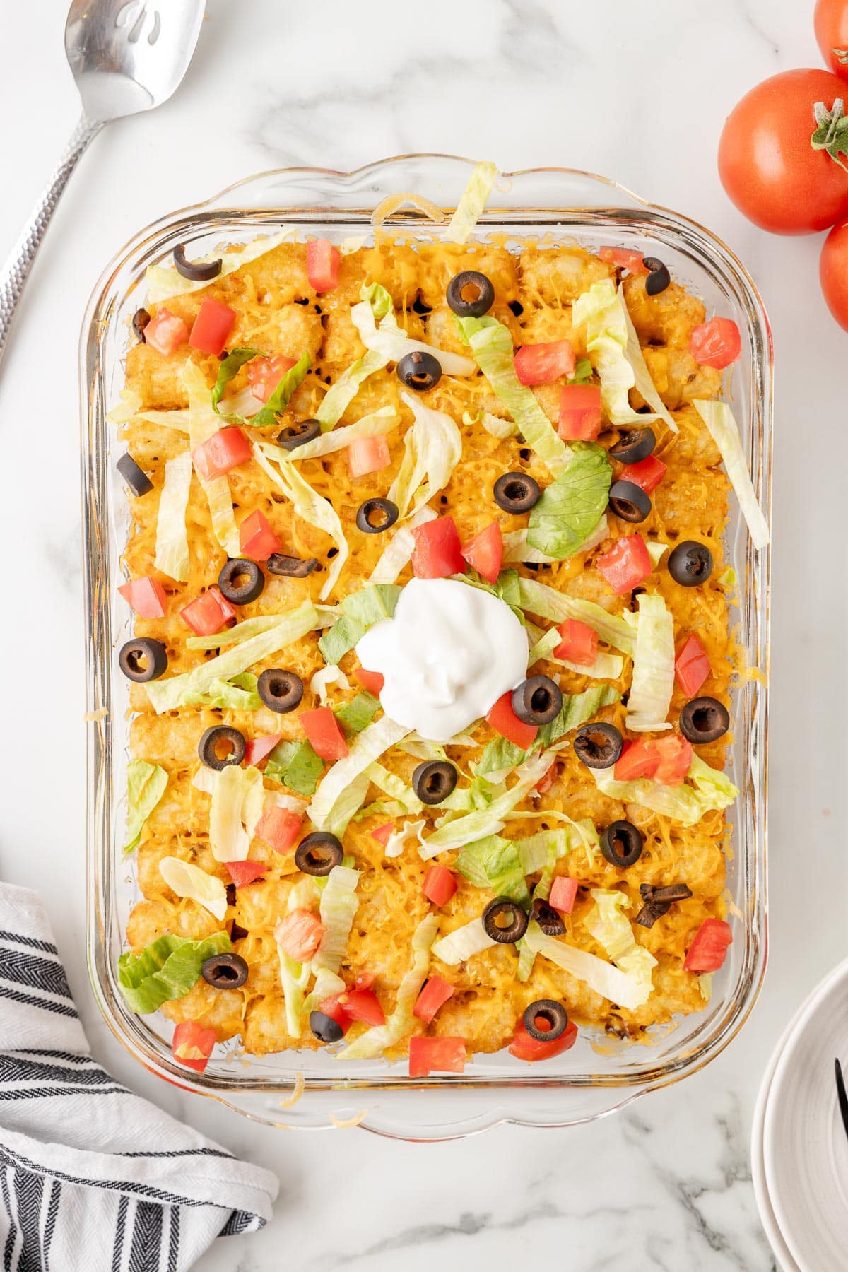 A finished taco tater tot casserole topped with cheese, sliced olives, diced tomatoes and shredded lettuce in a glass baking dish from above.