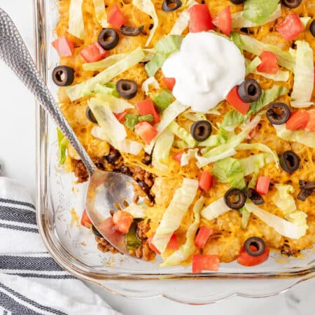 A glass dish with layered taco tater tot casserole topped with cheese, tomatoes, black olives, lettuce, and a dollop of sour cream while a spoon scoops a serving.