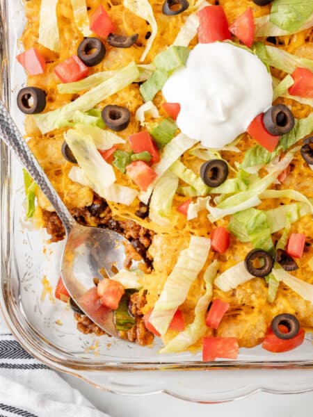 A glass dish with layered taco tater tot casserole topped with cheese, tomatoes, black olives, lettuce, and a dollop of sour cream while a spoon scoops a serving.