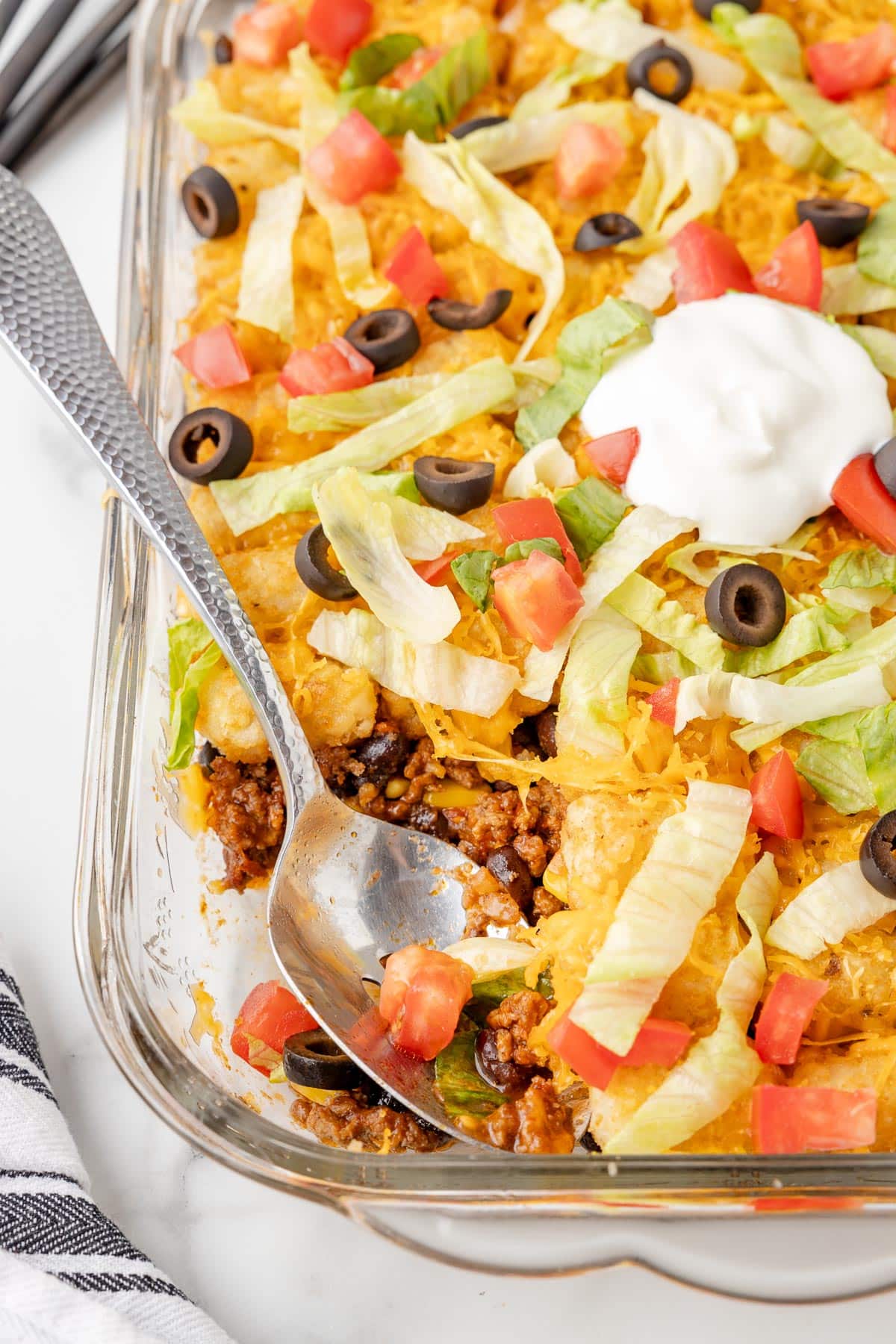 A casserole dish filled with a taco tater tot casserole topped with shredded lettuce, diced tomatoes, black olives, cheese, and a dollop of sour cream while a serving spoon scoops a portion.