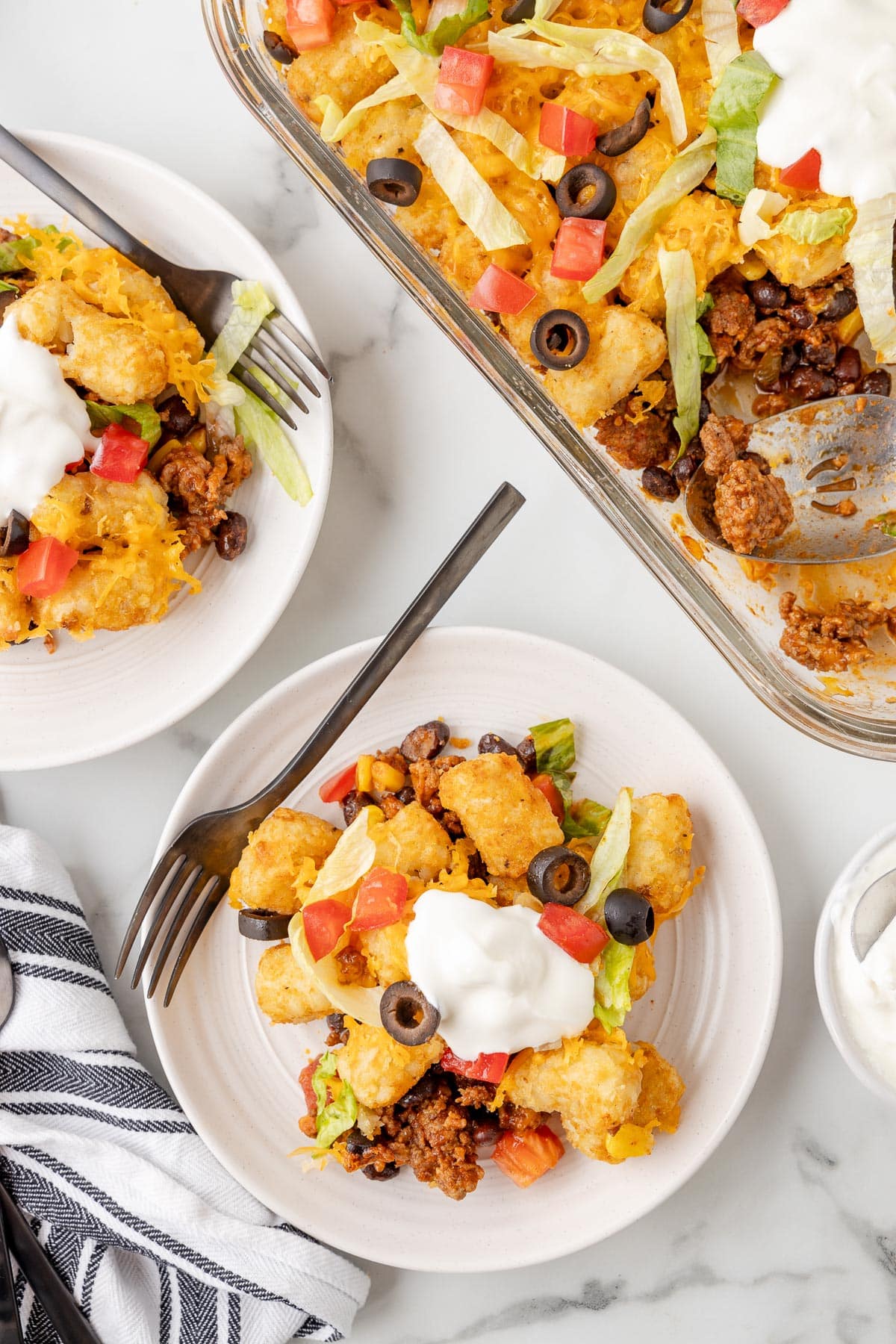 A dish filled with a serving of taco tater tot casserole next to a second plate with a serving and a full casserole pan with a scoop missing.