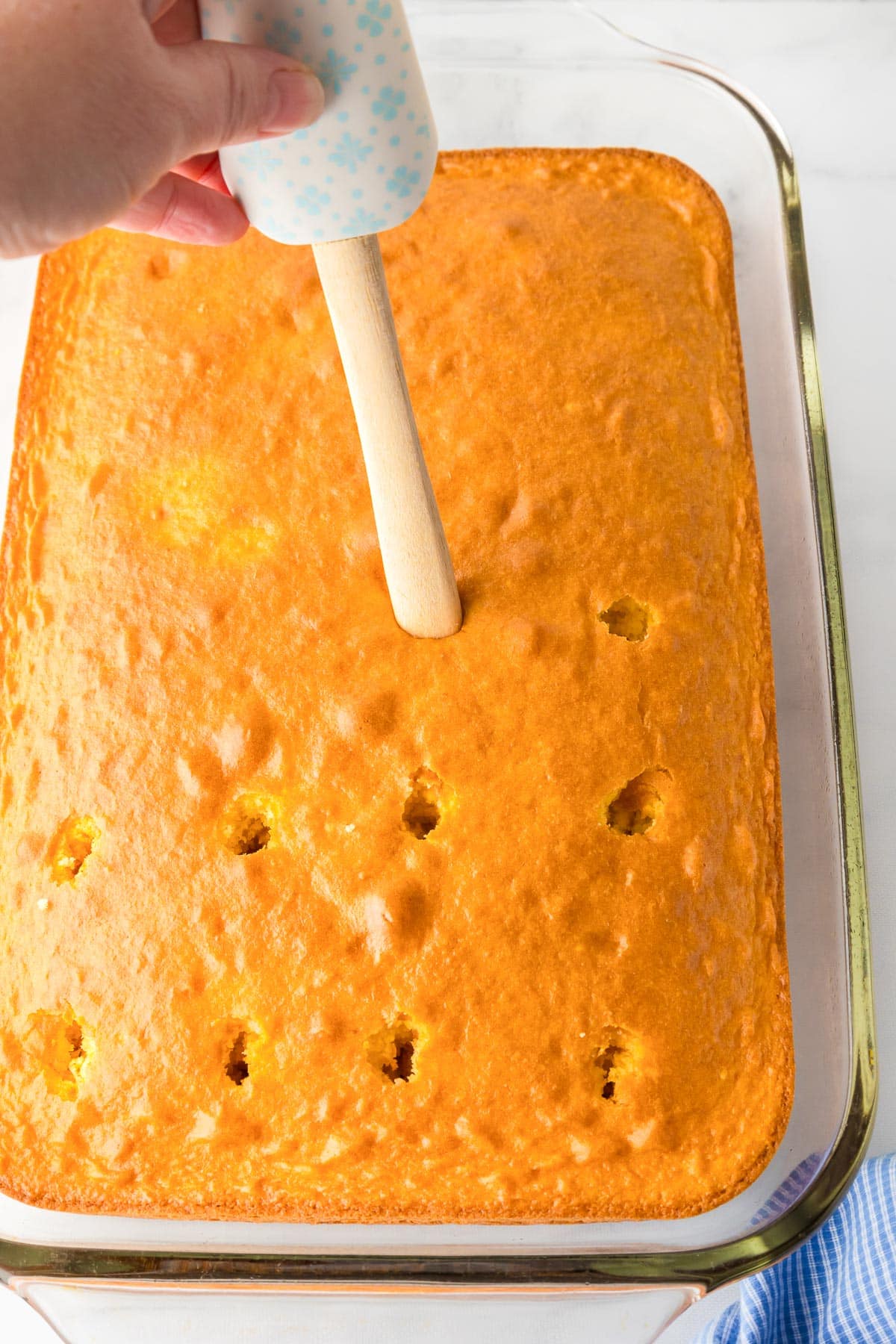 A hand uses the wooden handle of a spatula to poke round holes in a yellow cake in a rectangular baking dish.