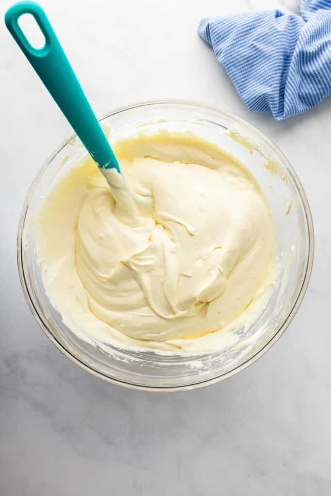 A glass bowl filled with creamy cream cheese frosting being mixed with a spatula for banana pudding poke cake.