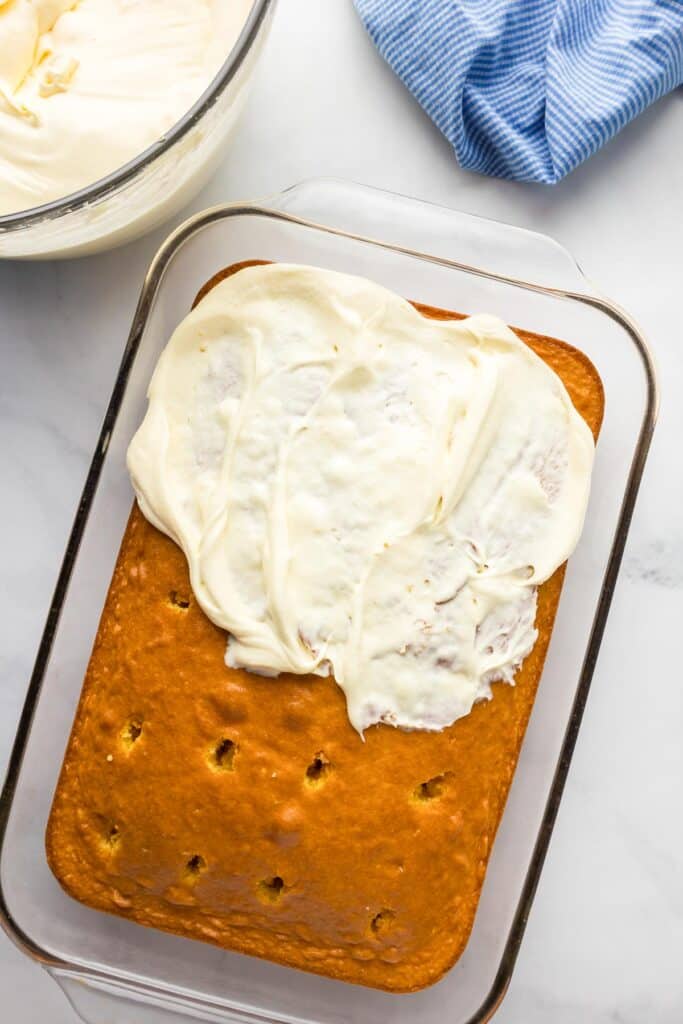 A rectangular yellow cake with holes in a glass casserole dish being frosted with a cream cheese mixture.