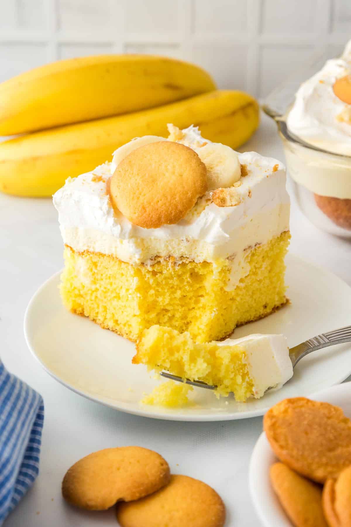 A slice of yellow banana pudding poke cake on a plate with whipped cream topping, vanilla wafers, and slices of banana while a fork cuts a bite.