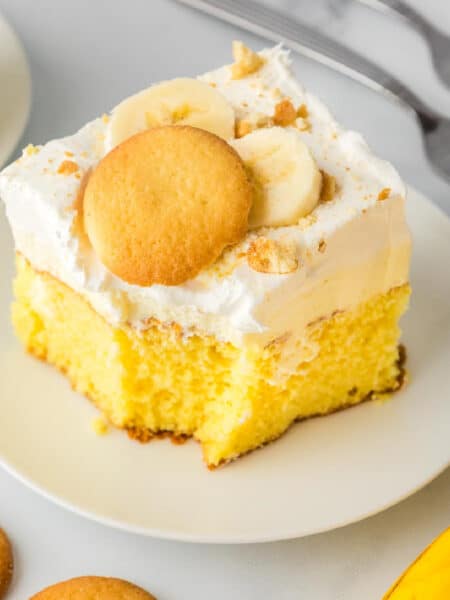 Square vies of a slice of yellow banana pudding cake topped with whipped cream frosting, banana slices, and a vanilla wafer cookie on a plate.