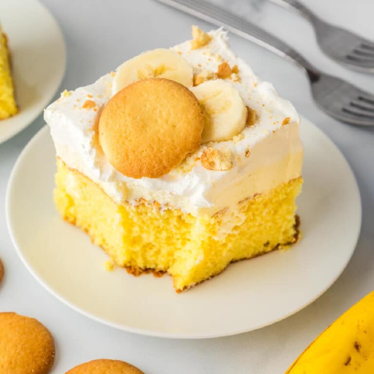 Square vies of a slice of yellow banana pudding cake topped with whipped cream frosting, banana slices, and a vanilla wafer cookie on a plate.