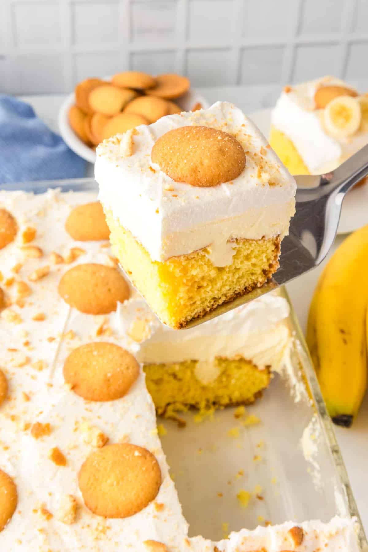 A slice of banana pudding cake topped with whipped cream frosting and a cookie is lifted from a baking dish with a spatula.