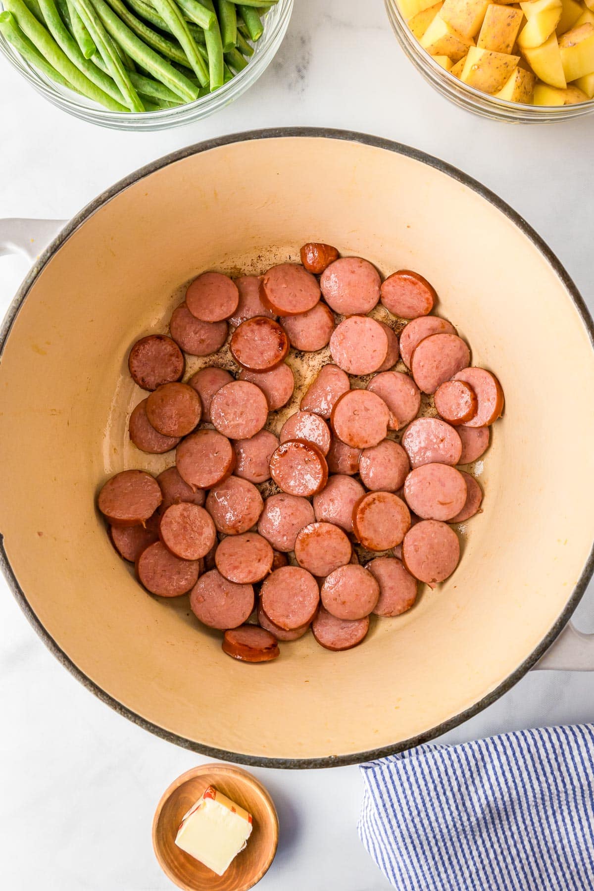 Slices of smoked sausage cooking in a large pot, with bowls of green beans, diced potatoes, and a small dish of butter nearby on a kitchen countertop.