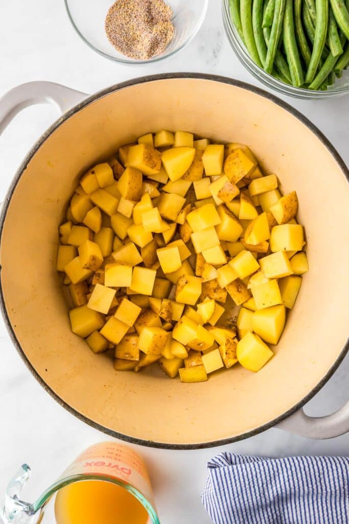 Chopped yellow potatoes in a pot before cooking, with bowls of seasoning, green beans and broth nearby.