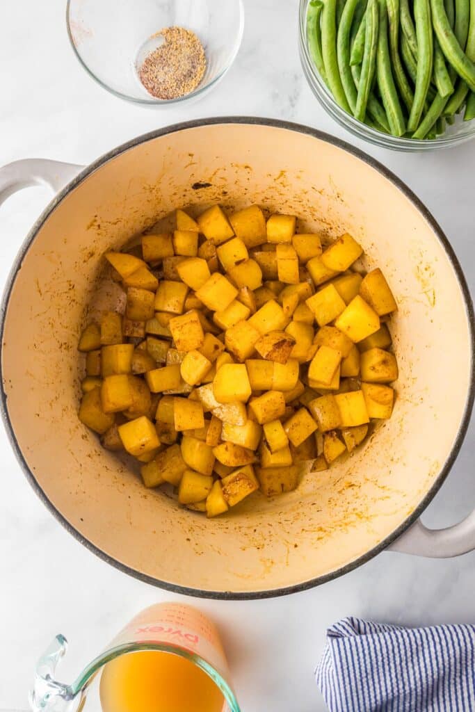 Cooked browned and cubed yellow potatoes seasoned in a large pot, surrounded by green beans, spices in a bowl, broth in a measuring cup, and a striped cloth.