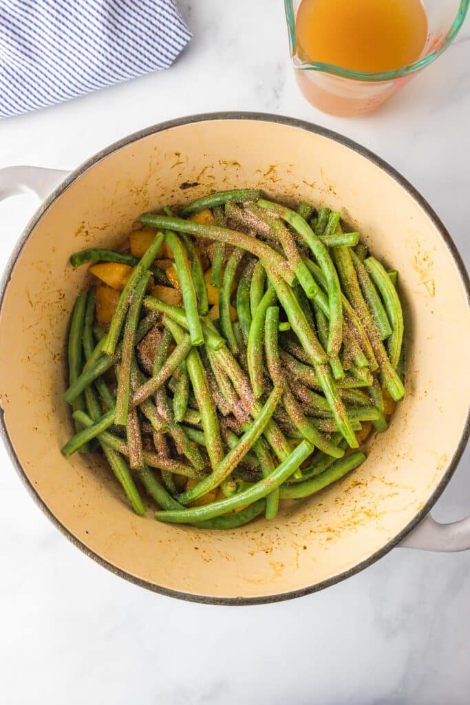 Uncooked green beans in a pot over cooked potatoes with spices on a counter with a measuring cup full of broth nearby on the counter for smoked sausage, green beans and potatoes.