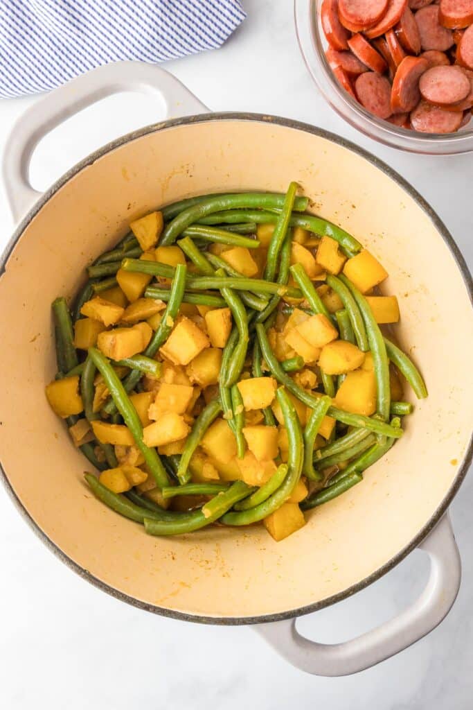 A pot with cooked green beans and potatoes with a bowl of cooked smoked sausage nearby.