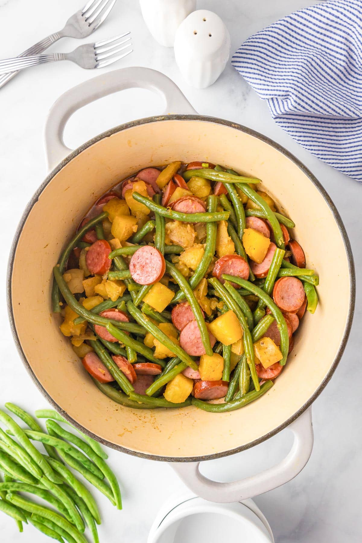 A pot filled with sliced smoked sausage, green beans, and chunks of yellow potatoes. Next to the pot are fresh green beans after cooking.