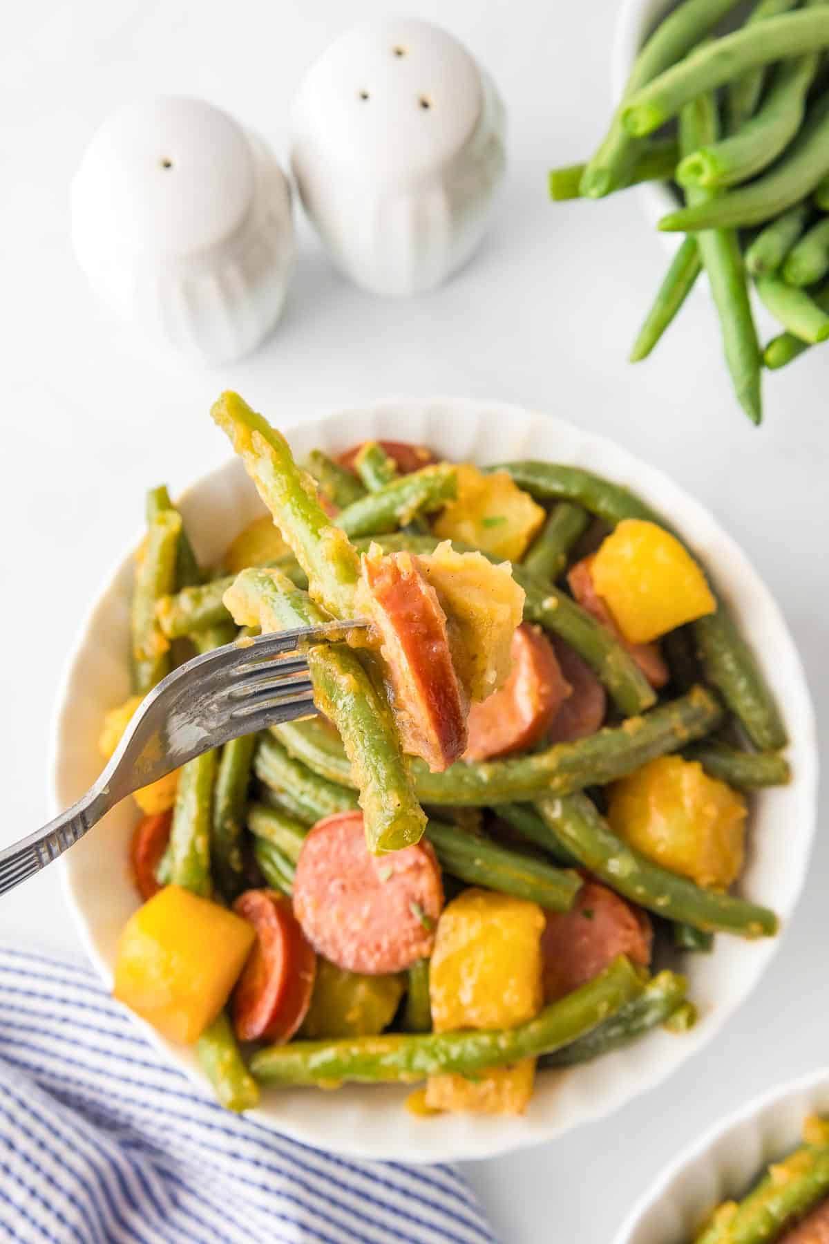 A bowl of cooked smoked sausage, green beans and potatoes with a fork holding a bite on a table.