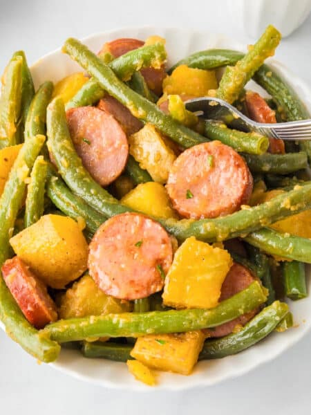 A bowl on a dinner table of green beans, sliced sausage, and chunks of yellow squash with a fork grabbing a bite.