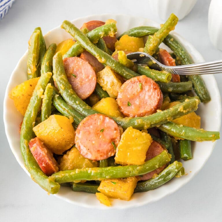 A bowl on a dinner table of green beans, sliced sausage, and chunks of yellow squash with a fork grabbing a bite.