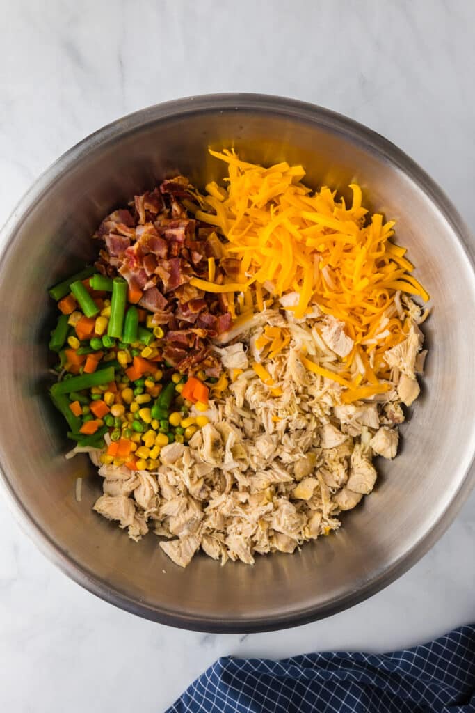 A bowl containing shredded chicken, grated cheese, bacon bits, and mixed vegetables on a marble surface.