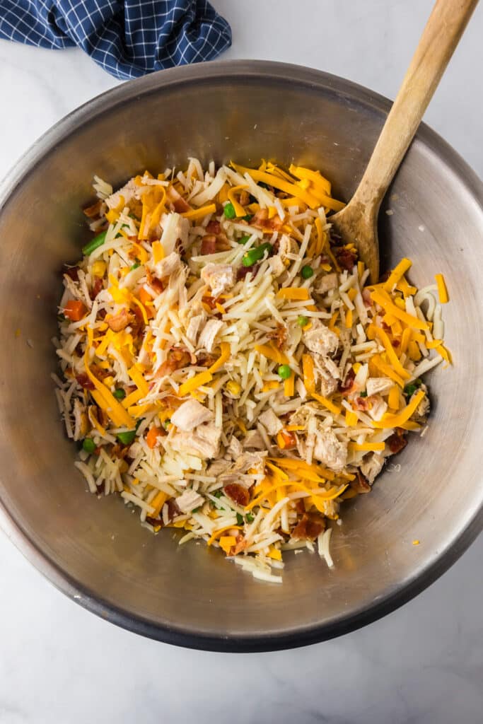 A bowl of shredded cheese, cooked chicken pieces, bacon pieces and chopped vegetables mixed with shredded hash browns with a wooden spoon.