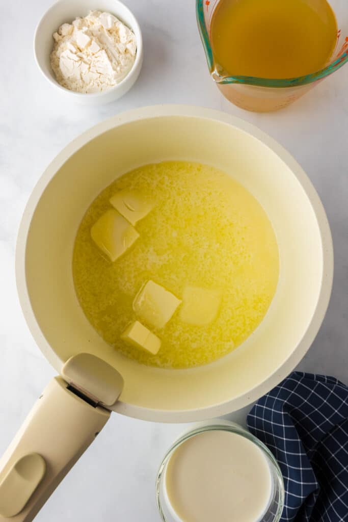 Butter melting in a saucepan on a marble surface, with bowls of flour, chicken broth and heavy cream nearby.