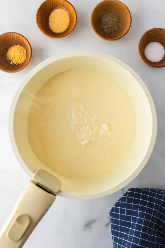 A smooth white sauce in a saucepan surrounded by wooden bowls containing different spices on a counter from overhead.