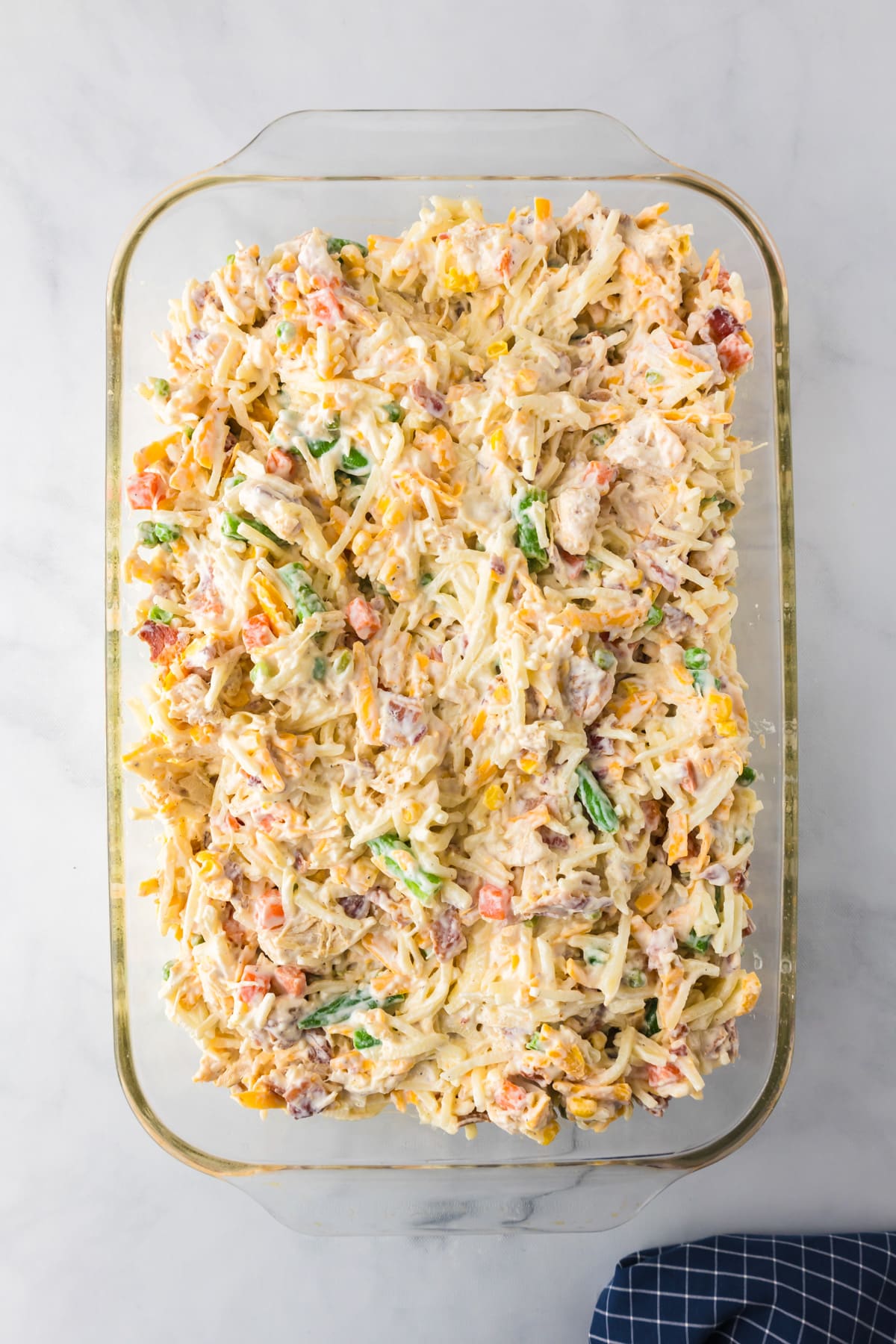 A glass baking dish filled with a hash brown casserole mixed with chicken, bacon, vegetables cheese, sour cream and white sauce placed on a countertop.