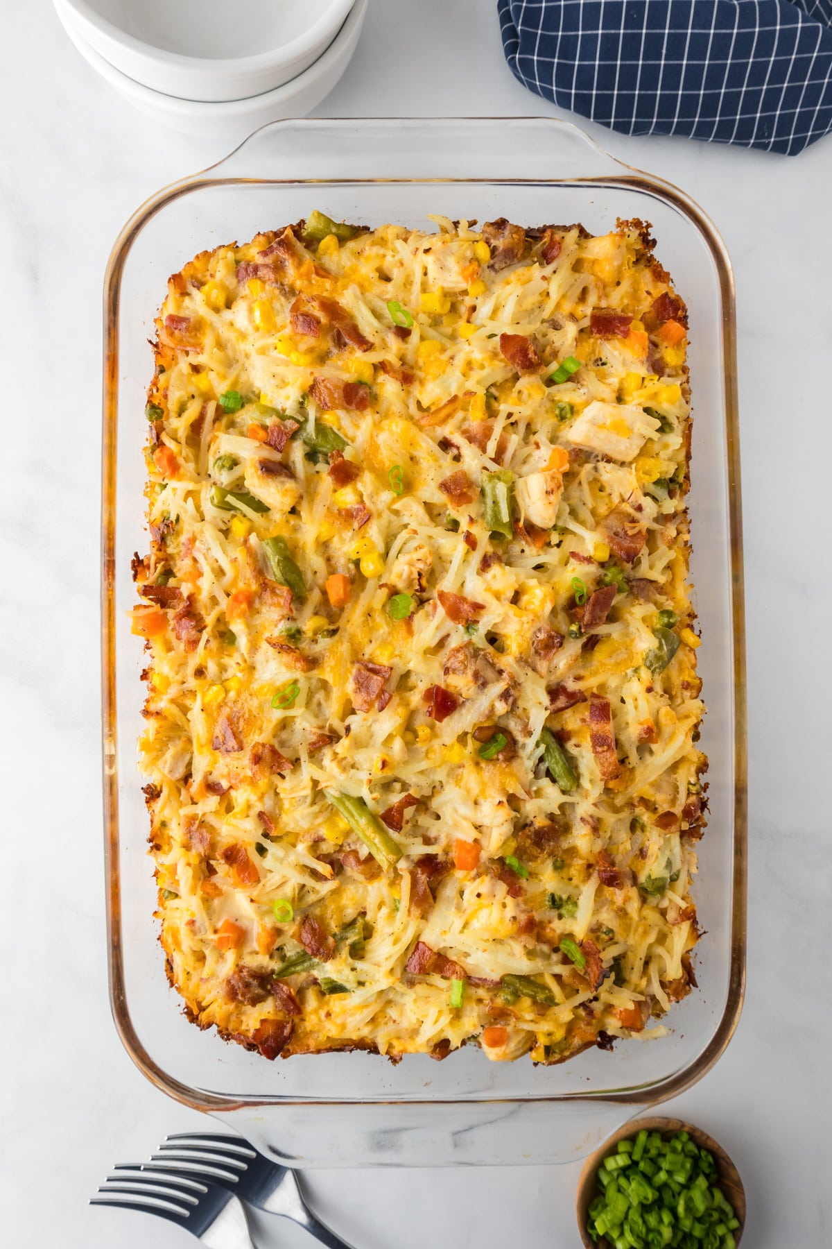 Baked casserole dish with golden brown hash browns, chicken, bacon, mixed vegetables, and melted cheese, placed on a countertop next to bowls and forks on a counter.