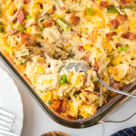 A square view of a baked chicken hashbrown casserole with a spoon scooping a mixture of potatoes, chicken, bacon and vegetables in a casserole pan.