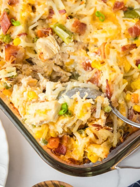 A square view of a baked chicken hashbrown casserole with a spoon scooping a mixture of potatoes, chicken, bacon and vegetables in a casserole pan.