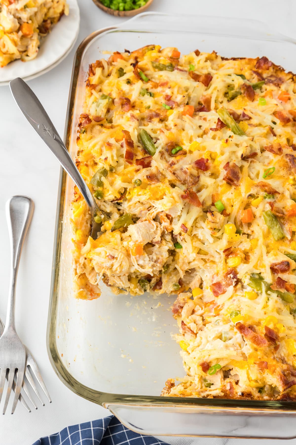 A baked casserole dish with a portion removed, showing mixed vegetables, shredded chicken, hash browns, and cheese. A serving spoon rests in the dish. Two forks are placed beside it.