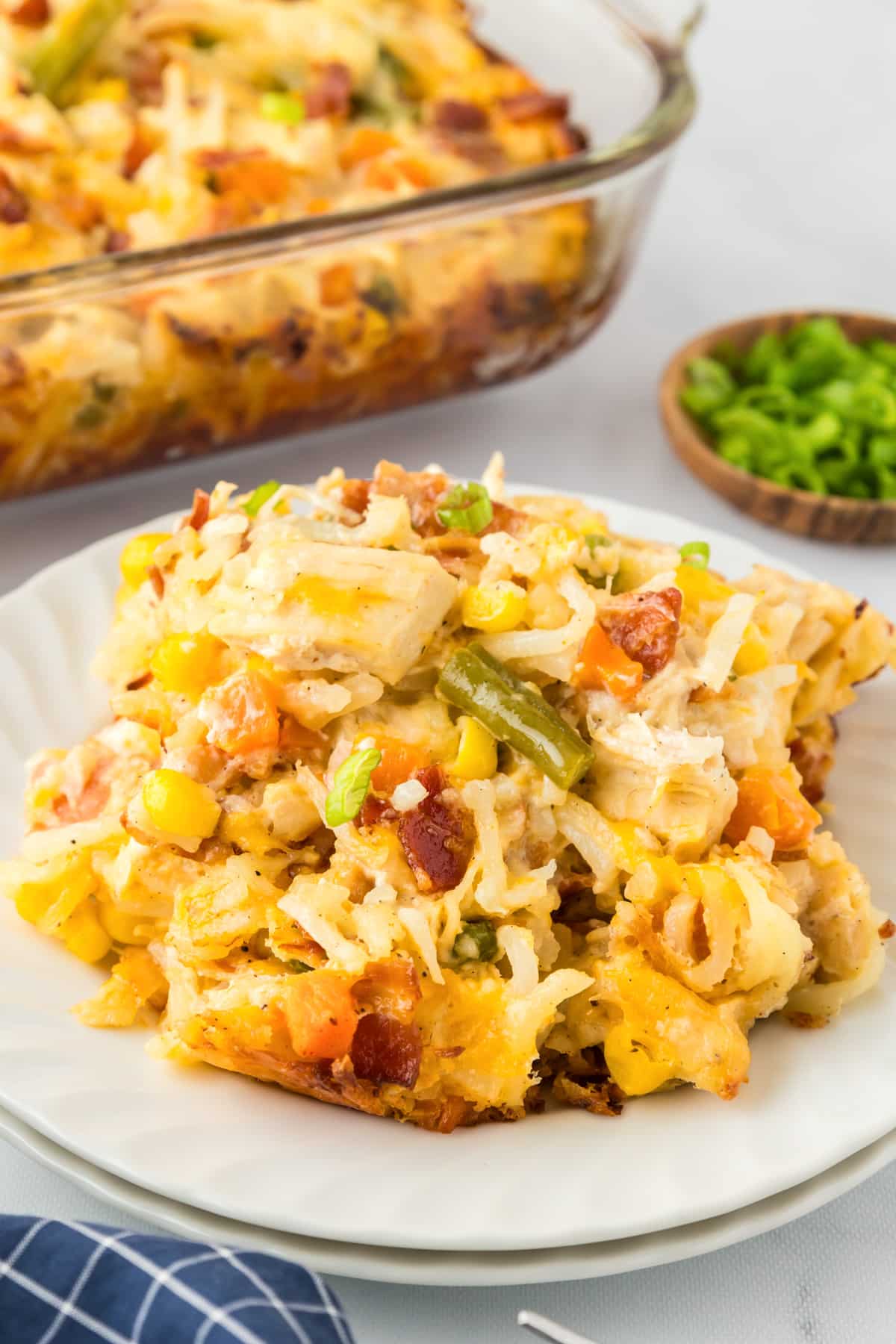 A serving of chicken hashbrown casserole with vegetables, cheese, chicken, vegetables and bacon on a white plate with a casserole dish full of more casserole in the background.