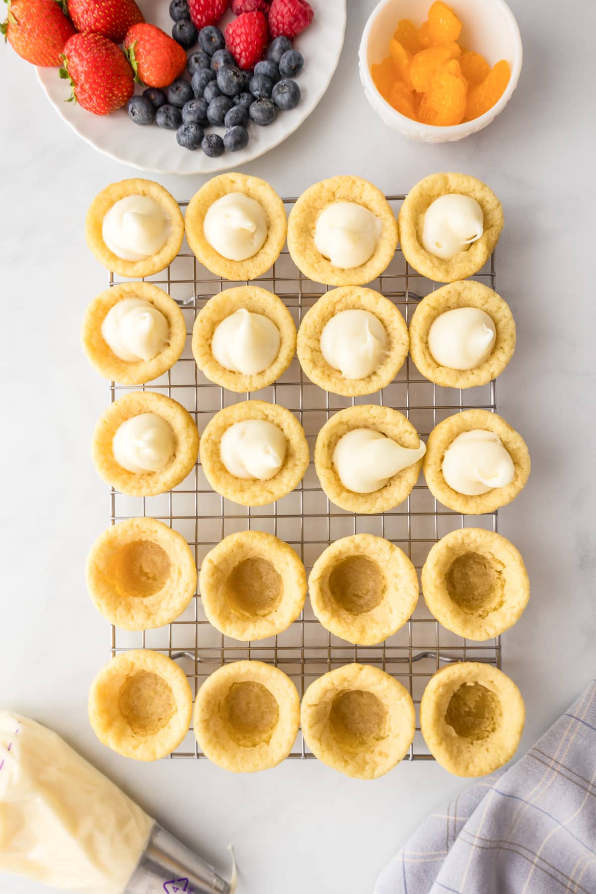 Sugar cookie cups on a cooling rack, half filled with the cream for fruit pizza frosting with fresh fruit on the counter nearby.