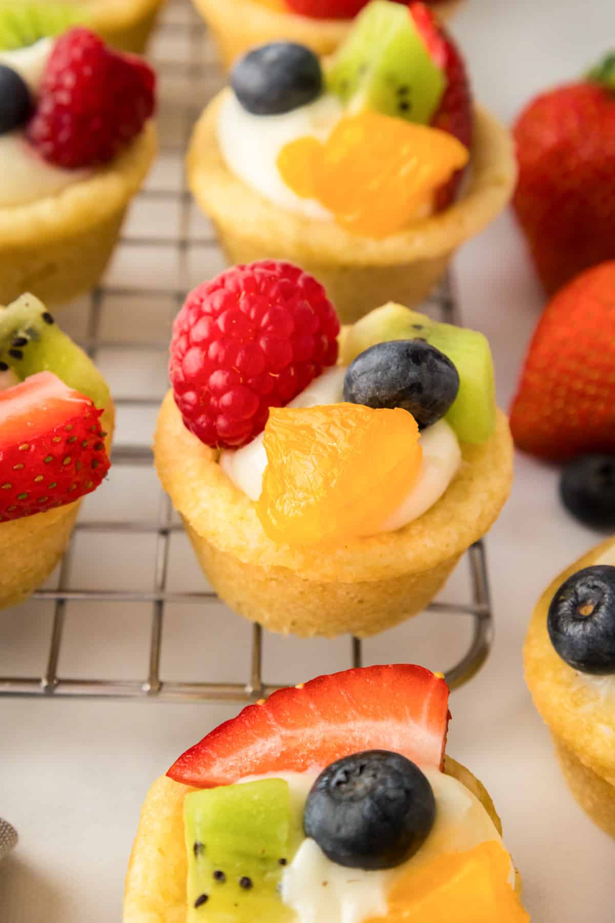 Fruit pizza cookie cups with cream filling, topped with fresh berries and other fruit on a cooling rack.