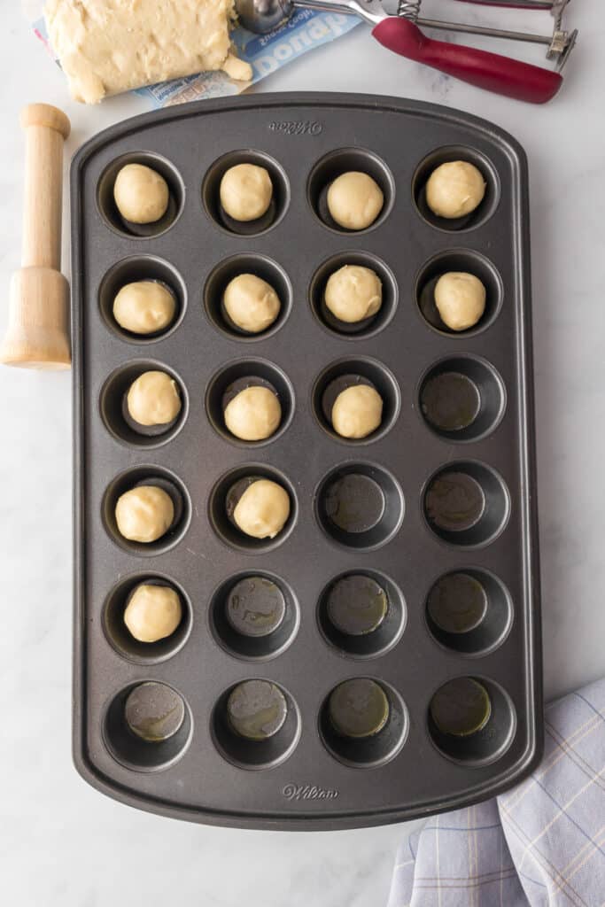 Mini muffin tin with 18 dough balls placed in each cup, alongside a rolling pin and dough piece on a marble countertop.