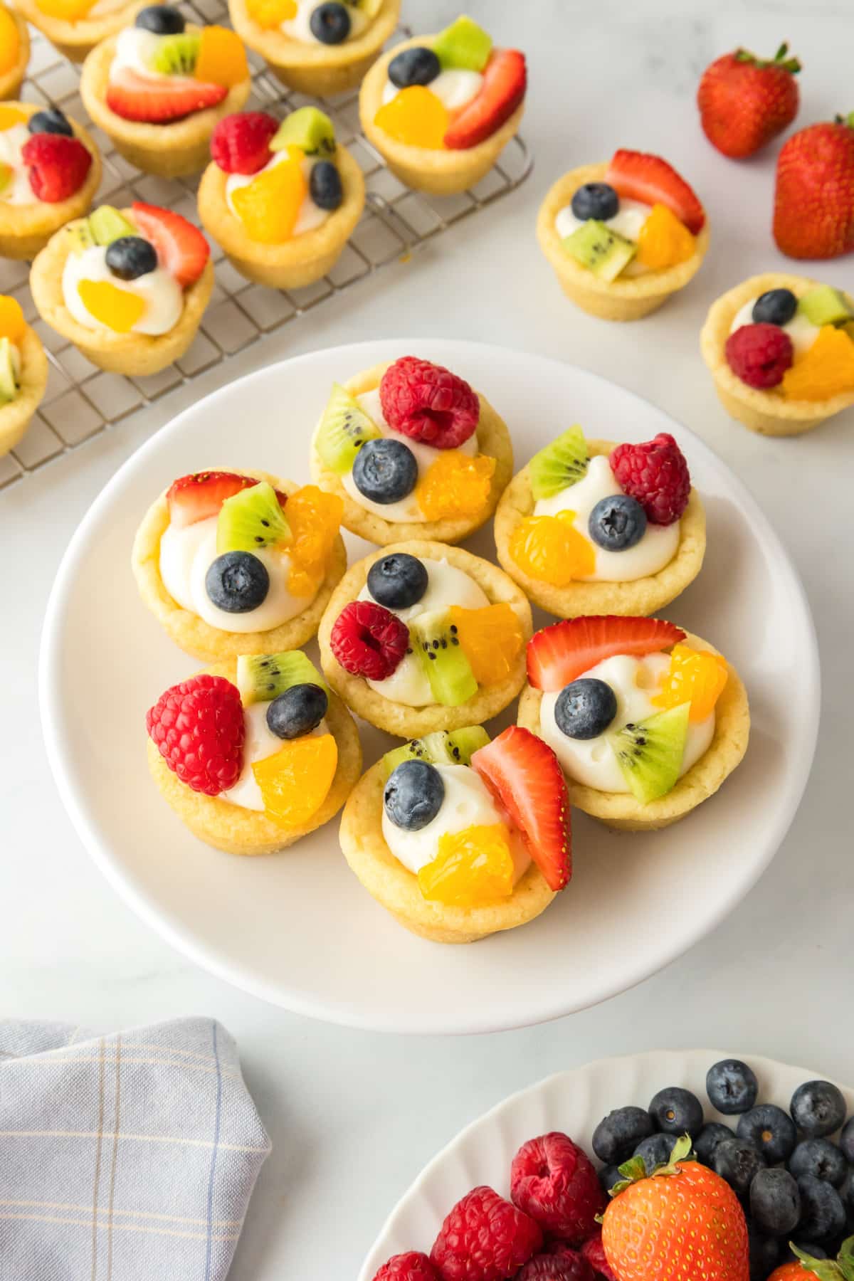 A white plate with fruit pizza sugar cookie bites topped with fresh fruit with more mini tarts in the background.