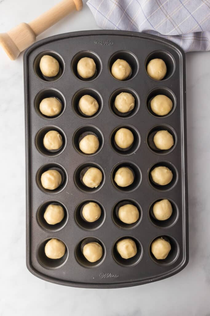 Mini muffin tray filled with round dough balls, placed on a marble surface with a striped cloth and wooden rolling pin nearby.