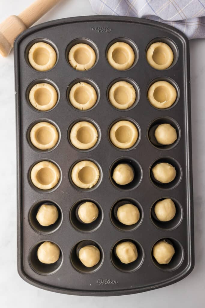 Mini muffin tin with dough balls and hollowed centers filled, ready for baking. A rolling pin and checkered cloth are partially visible.