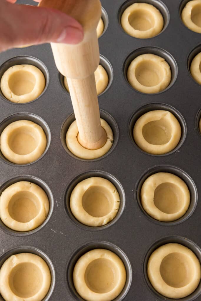 Hand pressing sugar cookie dough into a mini muffin tin using a wooden dowel, creating hollow centers in each cookie dough piece.