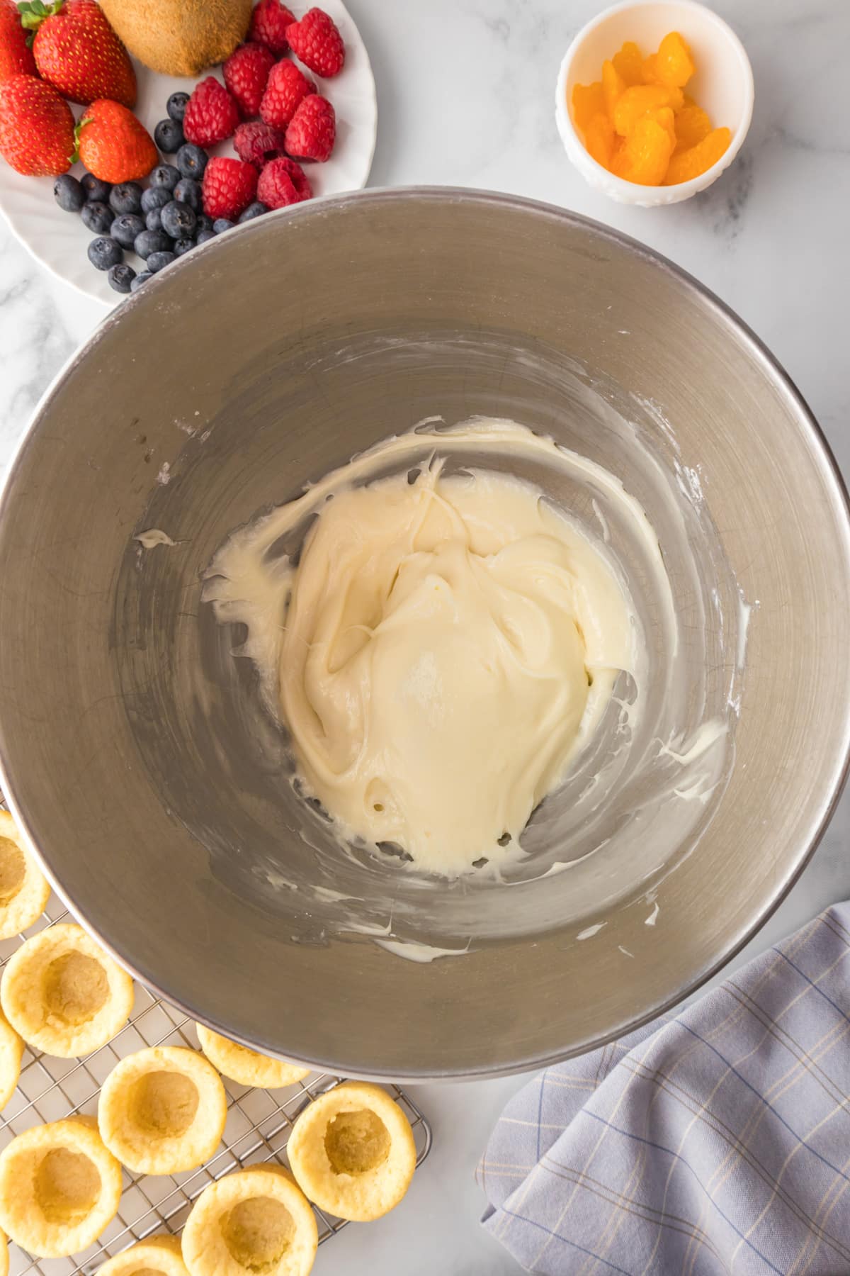 A mixing bowl with cream cheese frosting being mixed with baked cookie cups and fresh fruit nearby to assemble the fruit pizza cookie cups.