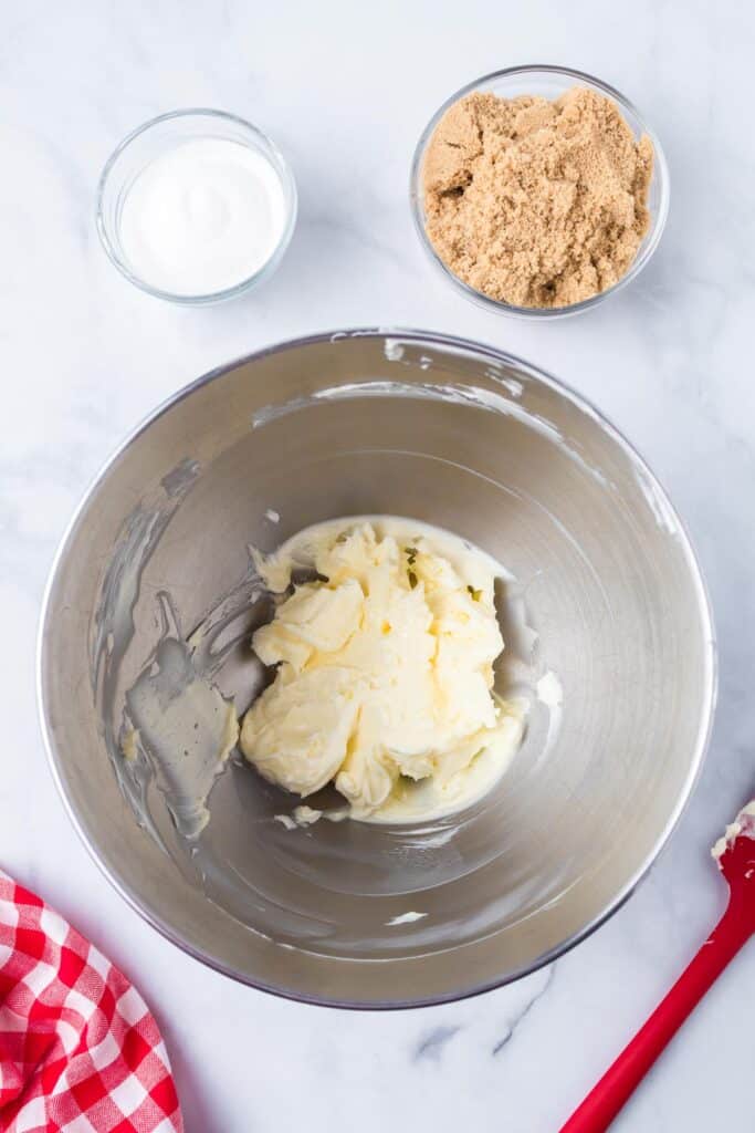 Mixing bowl with softened butter, surrounded by bowls of white sugar, brown sugar on a counter from above.