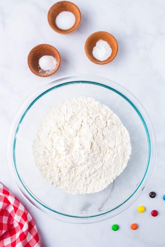 A mixing bowl with flour and dry ingredients in small bowls nearby to be mixed for M&M cookie bars.