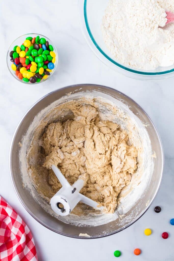 Cookie dough in a mixing bowl with flour being added to the wet ingredients with a small bowl of M&Ms nearby.