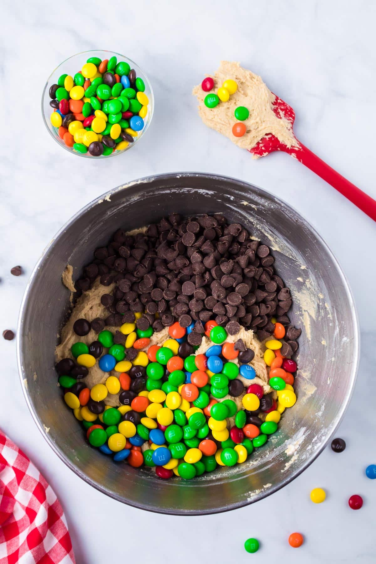 Mixing bowl with cookie dough, chocolate chips, and M&M candies being mixed together on a counter from above.