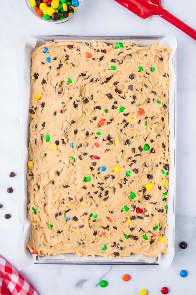 A baking tray with raw cookie dough pressed into a parchment lined pan mixed with M&MS and chocolate chips.